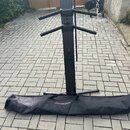 Černý kovový stojan na klavír s rozkládacími rameny. U základny je položen černý přepravní vak s bílým nápisem ULTIMATE SUPPORT. Povrch stojanu i vak…