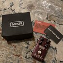 Na fotografii je tmavě červený efekt pedál s nápisy MXR, Super Badass a VARIAC FUZZ. Vedle pedálu je černá krabice s logem MXR, plastový sáček a dva…