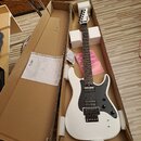 Bílá elektrická kytara v kartónovém přepravním obalu s polystyrenovými výplněmi. Na hlavě nástroje je viditelné logo Schecter, kytara má černý pickgu…