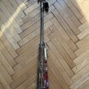 Fotografie zobrazuje kovový basový pedál v chromovaném provedení. Nástroj je položen na dřevěné podlaze. Na povrchu nejsou vidět žádné čitelné značky…
