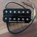 Černý humbucker s kovovými póly a bílým nápisem Seymour Duncan. Nástroj má připojený kabel s několika barevnými vodiči. Povrch je lesklý a vykazuje m…
