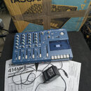 Modrý vícestopý magnetofon s nápisem TASCAM a označením PORTSTUDIO 414. Za přístrojem je umístěn originální karton s nápisy TASCAN, TASCAM a 4mkII. V…