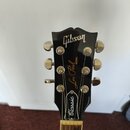 Černá hlava elektrické kytary se stálkovými ladičkami. Na povrchu je bílé logo Gibson a žlutý nápis Les Paul MODEL oraz bílý nápis Classic. Povrch je…