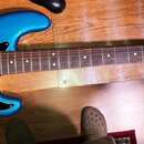 Modrá elektrická basová kytara s černým pickguardem a hnědým hmatníkem. Na hlavě nástroje je viditelné logo Fender a nápis PRECISION BASS. Nástroj vy…