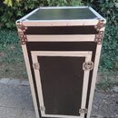 Černý transportní kufr s hliníkovými hranami a kovovými rohy. Přední strana obsahuje čtvercový dvířka s dvěma kovovými zámky. Povrch vykazuje mírné z…