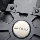 Detailní pohled na černý plastový prvek s kruhovým stříbrným logem ALESIS v centru. Povrch plastu vykazuje drobné škrábance a prach. Nad logem je vid…