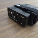 Černý obdélníkový elektronický přístroj s připevněným černým popruhem. Na zadní straně jsou viditelné konektory a nápisy: PED 1, 9V, in, out, PED 2 a…