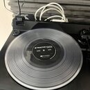Černý turntable s položeným průsvitným vinylovým diskem. Na štítku disku je bílý nápis TRAKTOR SCRATCH a logo Native Instruments. Pod ním je uveden t…