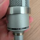 Stříbrný kondenzátorový mikrofon s kovovou síťovkou. Na těle je černý pruh s bílým nápisem MICROPHONE TLM 102 CARDIOID. Povrch je matně stříbrný, v h…