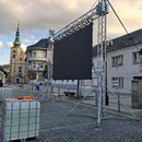 Fotografie zachicuje velkou černou LED stěnu namontovanou na hliníkovém trussovém systému. Konstrukce je zajištěna modrými napínacími popruhy a stojí…