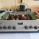 Stříbrný audio zesilovač v kovovém šasi s otevřeným horním krytem, uvnitř kterého jsou vidět dva velké transformátory a elektronické součástky. Na př…