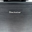 Detailní pohled na černý přední textilní kryt kytarového komba. Uprostřed je umístěn obdélníkový štítek s bílým nápisem Blackstar. Povrch textilie i…