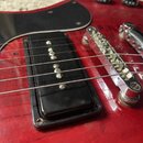 Detail červené elektrické kytary s černým pickguardem. Je vidět jeden černý humbucker snímač a chromovaný kobylník s připojenými strunami. Povrch kyt…
