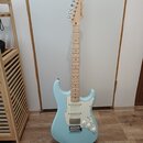 Světle modrá elektrická kytara s bílým pickguardem a javorovým hnedým hmatníkem. Na hlavě je viditelné logo "Wilkinson". Nástroj je umístěn na černém…