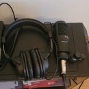 Na fotografii jsou černá uzavřená sluchátka a černý kondenzátorový mikrofon s kovovou mřížkou, umístěné na černém povrchu. Na těle mikrofonu je vidit…