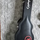 Černý tvrdý kufr na kytaru s rukojetí. Na horní části je bílý nápis Epiphone a v dolní části červený kruhový symbol s postavou zasaženou bleskem. Pov…