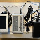 Na bílém kartonu s nápisem Touch Monitor TM3 je umístěn stříbrný dotykový displej s černým obrazovkou a logem RTW. Vedle něj je stříbrná perforovaná…