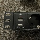 Černá zásuvková lišta s dvěma USB porty. Nad porty je bílé symbolické znázornění USB a červená kontrolní LED dioda. Dole je bílý nápis 5V == i 1A. Ve…
