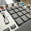 Bílo-šedý sampler s mřížkou šestiдесяti čtyř šedých gumových padů. Na horní části je nápis MPC SAMPLE, displej a několik ovládacích knoflíků a tlačít…