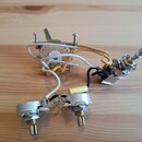 Fotografie zobrazuje elektroniku z elektrokitary, která obsahuje dva potenciometry a jeden přepínač. Komponenty jsou propojeny bílými a žlutými vodič…