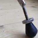 Detail kovové nohy s nastavitelnou výškou. Noha končí závitovým šroubem, na kterém je stříbrná podložka a černá gumová patka. Povrch kovu vykazuje zn…
