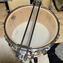 Fotografie zachycuje snare drum s napnutým kovovým sítkem (snares) přes bílý kožený Membrane. Na horním dřevěném ringu je černý nápis „REMO Ambassado…