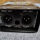 Zadní strana černého kovového zařízení s dvěma XLR konektory. Nad konektory jsou nápisy RIGHT OUT a LEFT OUT, pod nimi MIC LEVEL 600Ω. Na levém boku…