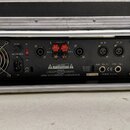Zadní strana černého rackového zesilovače v transportním kufru. Na panelu jsou nápisy FUSE, AC 200-240V, - BRIDGE +, OUT B, OUT A, IN B, IN A, XLR Pi…