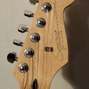 Fotografie zachycuje hlavu kytary z přírodního světlého dřeva s chromovanými ladičkami. Na povrchu je černý nápis Squier BY FENDER STRATOCASTER a v h…