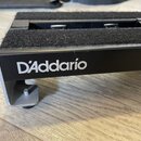 Černý kovový stojan s vrchní částí potaženou šedým textilem. Na boční straně je bílý nápis D'Addario. Povrch textilu vykazuje drobné nečistoty a prac…