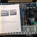 Míchací pult značky Behringer s nápisem XENYX QX1832USB. Nástroj je umístěn v přepravním kufru, vedle něj leží černý kabel a bílý papírový návod s ná…