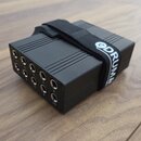 Černý obdélníkový box s žebrovaným horním povrchem a řadou deseti vstupních konektorů. Na boku jsou pod konektory viditelna čísla 1 až 10. Box je omo…