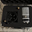 Kondenzátorový mikrofon stříbrné barvy s mřížkou a černý popfilter jsou umístěny v černé polstrované přepravní kufříku. Na těle mikrofonu je zlaté lo…