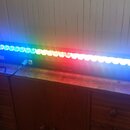 Černá lineární LED lampa s řadou svítících diod v barevněm přechodu od modré přes zelenou a červenou zpět k modré. Na předním panelu je digitální dis…