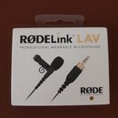 Fotografie znázorňuje bílé originální balení mikrofonu. Na krabici je uveden nápis RØDELink LAV a text PROFESSIONAL WEARABLE MICROPHONE, v pravém dol…