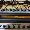 Detailní pohled na ovládací panel kontroleru s řadou posuvníků a knoflíků. Oranžový displej zobrazuje text „M-Audio Control Surface is in Logic Mode“…
