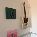 Elektrická kytara s krémovým povrchem těla a hnědým hmatníkem, zavěšená na bílém držáku na zdi. Kytara má tmavý pickguard a stříbrné kovové části. Na…