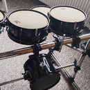Sada černých bubnů na chromovaném rackovém systému. Na blanách jsou vidět loga značky REMO. Nástroje vykazují běžné známky používání, povrch je leskl…