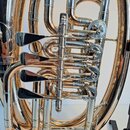 Detailní záběr na ventilový mechanismus mosazného nástroje v zlatém lesklém povrchu s chromovanými prvky. Jsou vidět čtyři ventilové písty s černými…