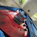 Elektrická kytara s červeným mramorovaným povrchem v modrém polstrovaném kufru. Nástroj má dva humbuckery, jeden přepínač a tři ovládací knoflíky. Na…