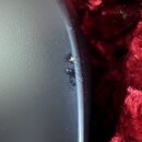 Detailní záběr na černý, hladký povrch hudebního nástroje nebo příslušenství. Na hraně je viditelné odřápání laku, pod kterým prosvítá světlý materiá…
