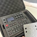 Černý drum computer a syntetizátor značky Elektron s nápisem Syntakt a doplňkem 12 Track Drum Computer & Synthesizer. Zařízení je umístěno v černé po…
