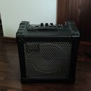 Černý kombozesilovač s kovovou ochrannou mřížkou a plastovými hranami. Na mřížce je umístěno logo a nápis Roland. Horní panel obsahuje ovládací knofl…