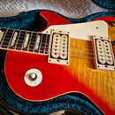 Detail elektrické kytary s červeno-žlutým sunburst povrchem v modrém pouzdře. Nástroj má dva krémové humbuckery, krémový pickguard a bílý oválný knof…