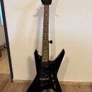 Černá elektrická kytara s ostře hranatým korpusem umístěná na stojanu. Na hlavě hmatníku je bílé logo Jackson. Nástroj má dva humbuckery, z nichž na…