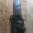 Černý polštovaný pouzdro (gig bag) na kytaru s bílým lemováním po okrajích. Má zipové hlavní oddíly a jednu menší zipovou kapsu na přední straně. Pov…