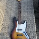 Čtyřstrunná elektrická basová kytara s tělem v provedení sunburst a bílým pickguardem. Na hlavě hředu je viditelné logo Fender. Nástroj vykazuje znám…