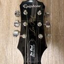 Černá hlava elektrické kytary s nápisem Epiphone v horní části. Uprostřed je zlatý nápis Les Paul a pod ním text MODEL. Ve spodní části hlavy je bílý…