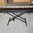 Černý digitální piano umístěný na černém X-tvarovém stojanu. Na přední straně nástroje jsou bílé nápisy Roland a FP-50. Povrch instrumentu vypadá čis…