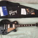 Černá elektrická kytara s hnědým pickguardem a dvěma humbuckery. Na hlavě nástroje je bílé logo Gibson. Vedle kytary je černý obal s bílým nápisem Gi…